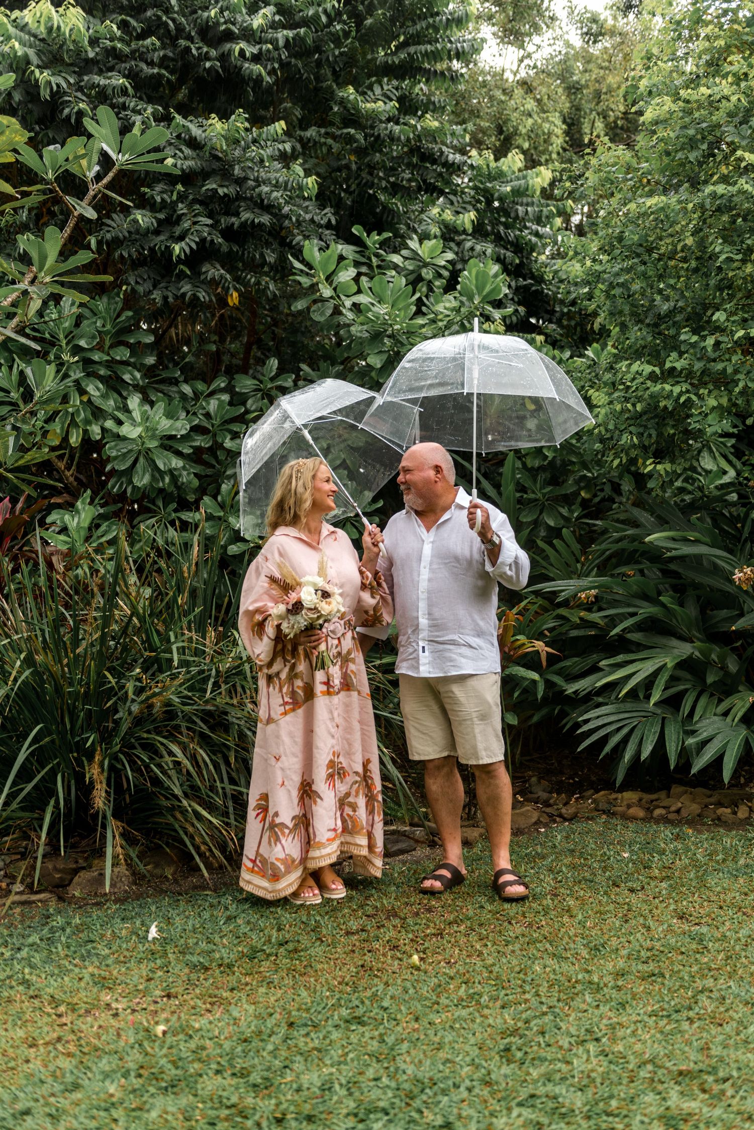 Mature couple sharing a smile under clear bubble umbrellas in a lush tropical garden setting.