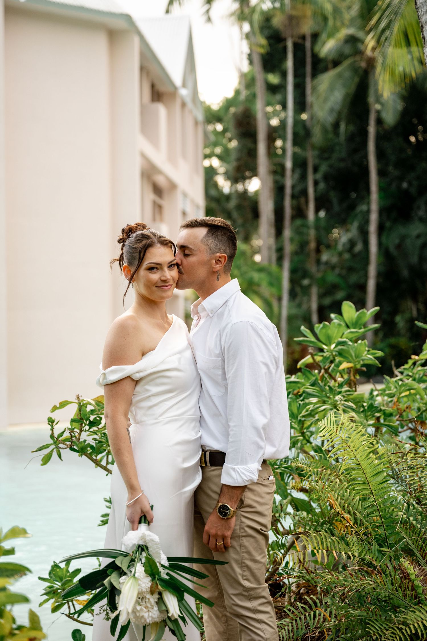 Groom kissing bride on the cheek beside a tropical resort pool with lush green surroundings at Sheraton Grand Mirage Port Douglas.