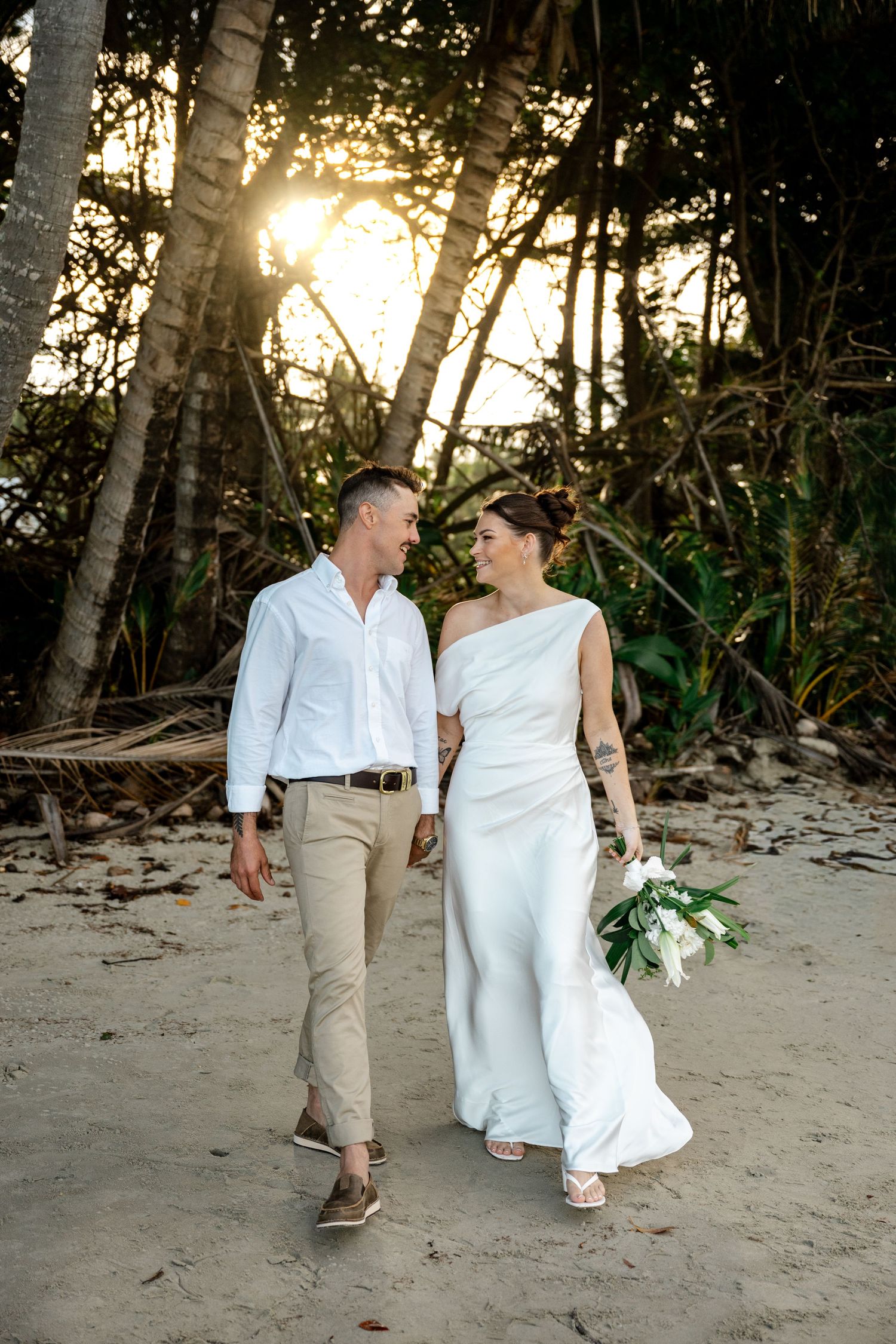 Newlyweds walk hand in hand on a sandy tropical beach at golden hour, bride holding white bouquet at Four Mile Beach, Port Douglas.