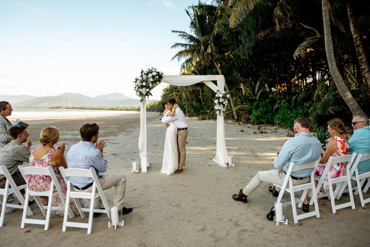 Bride and groom sharing their first kiss at a beach wedding ceremony with guests seated on white chairs at Four Mile Beach, Port Douglas.