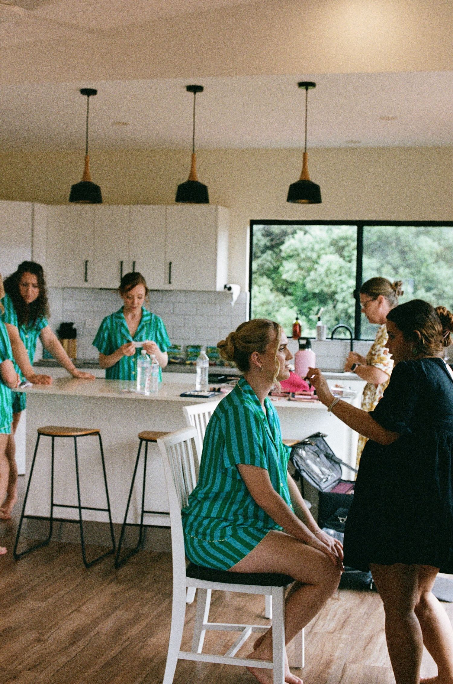 Bridesmaids wearing teal striped pajamas get ready together in a bright white kitchen.