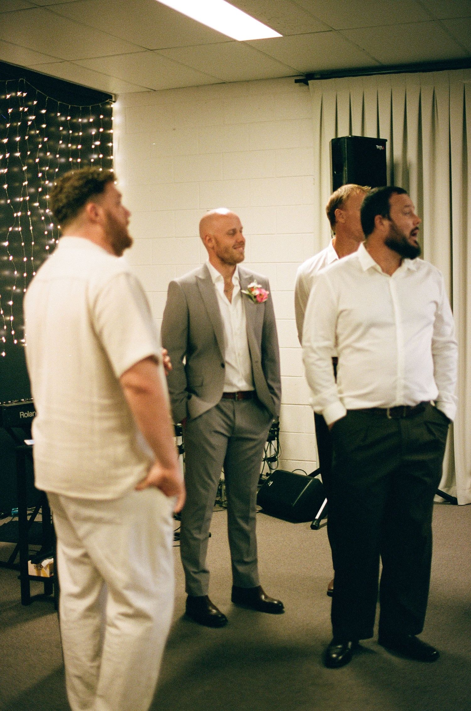 Groom in gray suit with pink boutonniere stands with groomsmen before wedding ceremony near fairy light backdrop.