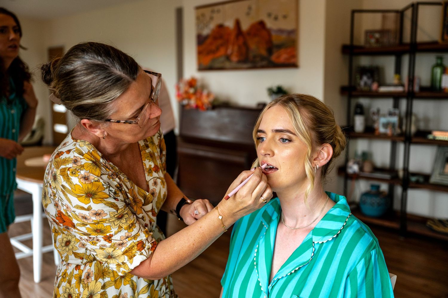 Makeup artist carefully applying foundation to blonde bride in teal pajamas in bright home interior.