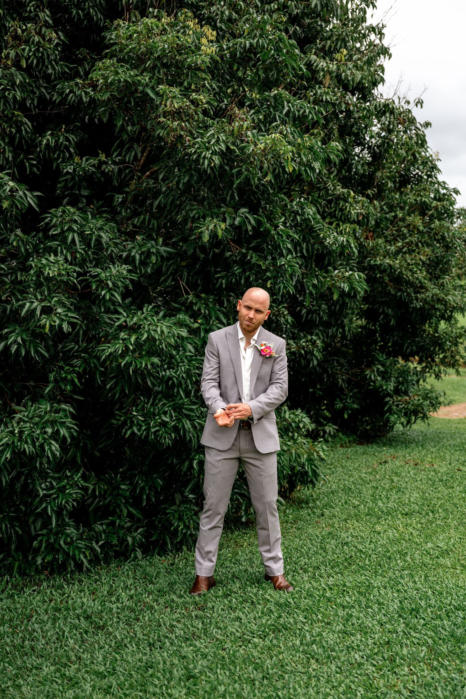 Bald groom in grey suit with pink boutonniere posing with clasped hands before green trees.