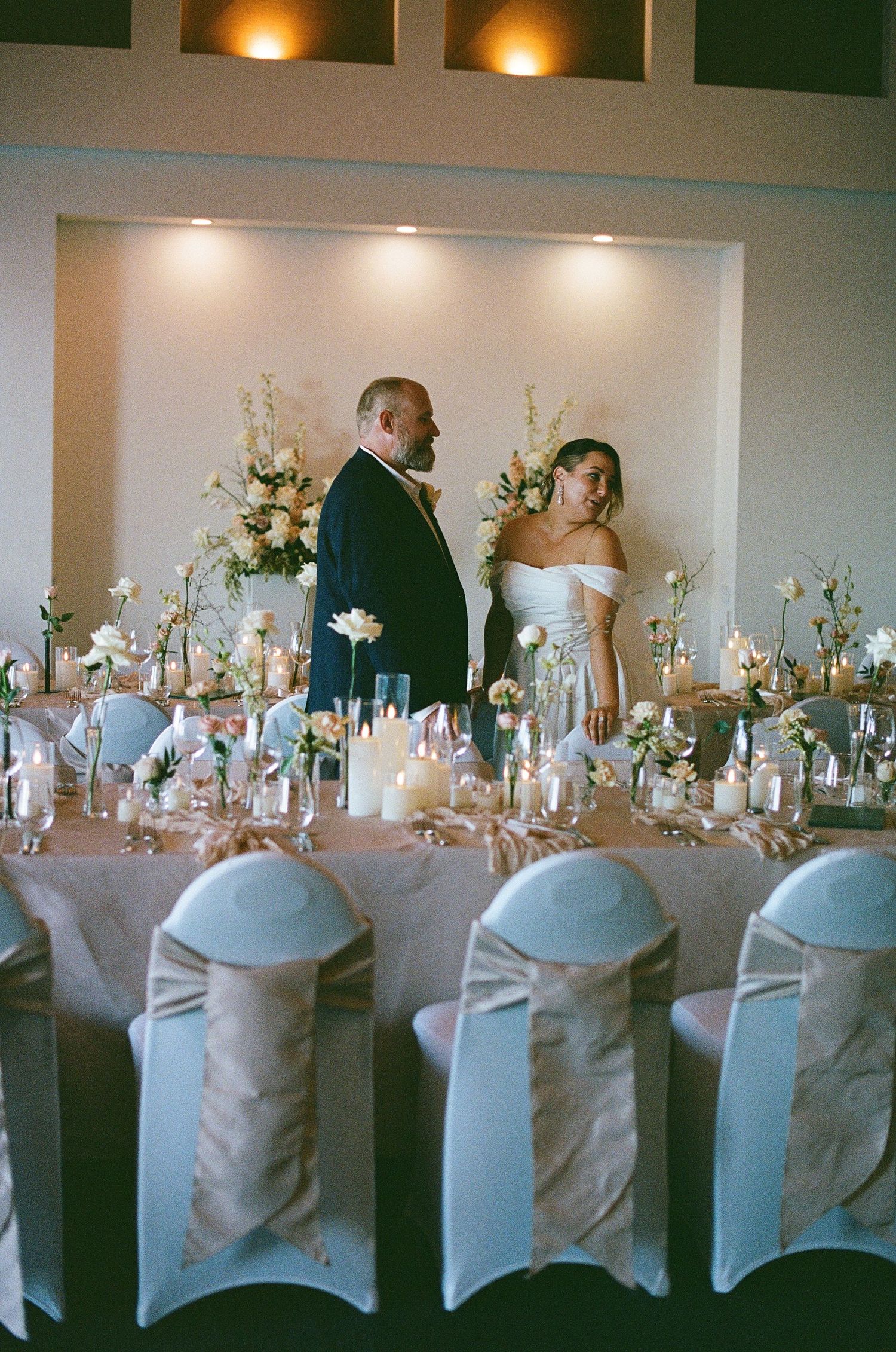 Bride and groom standing at elegantly decorated wedding reception head table with candles and floral arrangements.