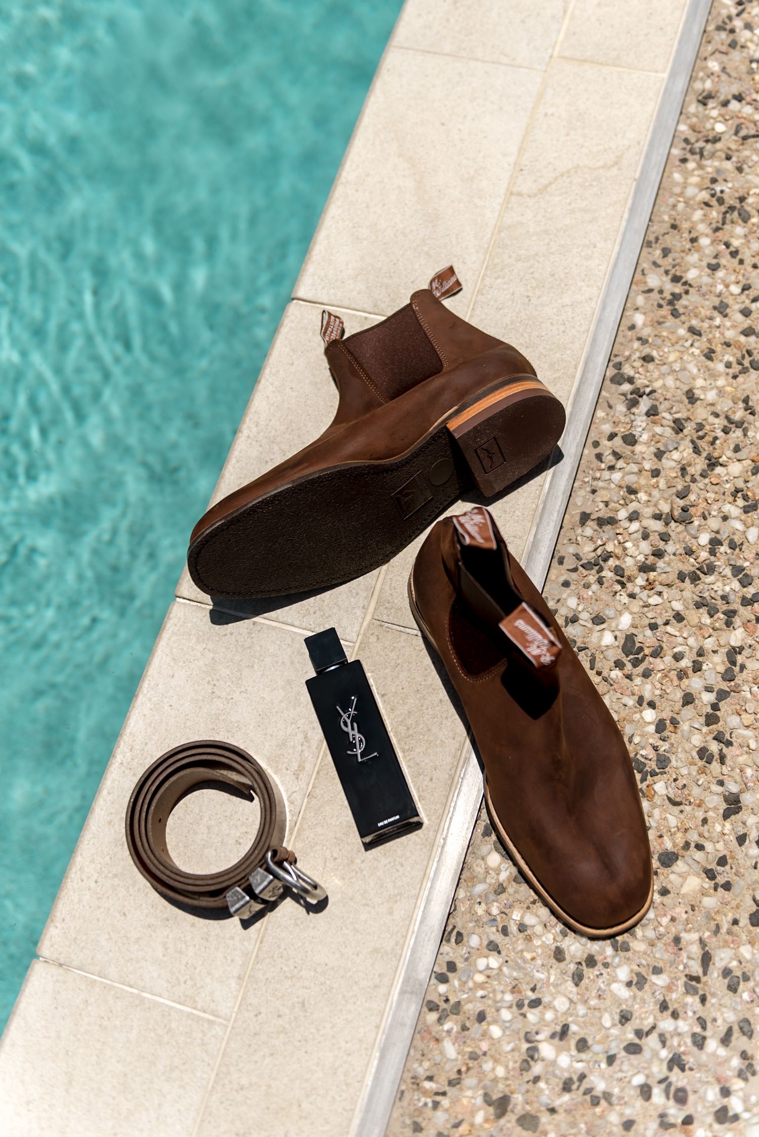 Brown suede Chelsea boots with YSL cologne and leather belt displayed poolside on marble surface.