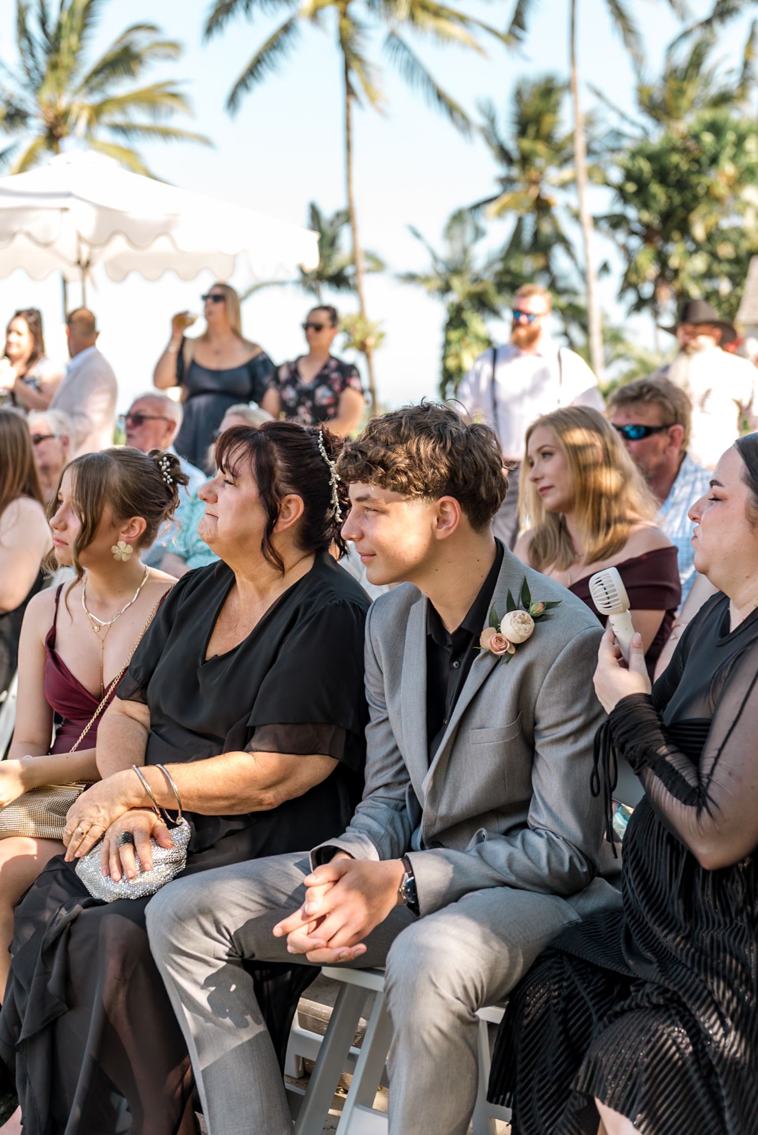 Young wedding guest in gray suit smiles warmly while seated among family during sunny outdoor tropical ceremony.