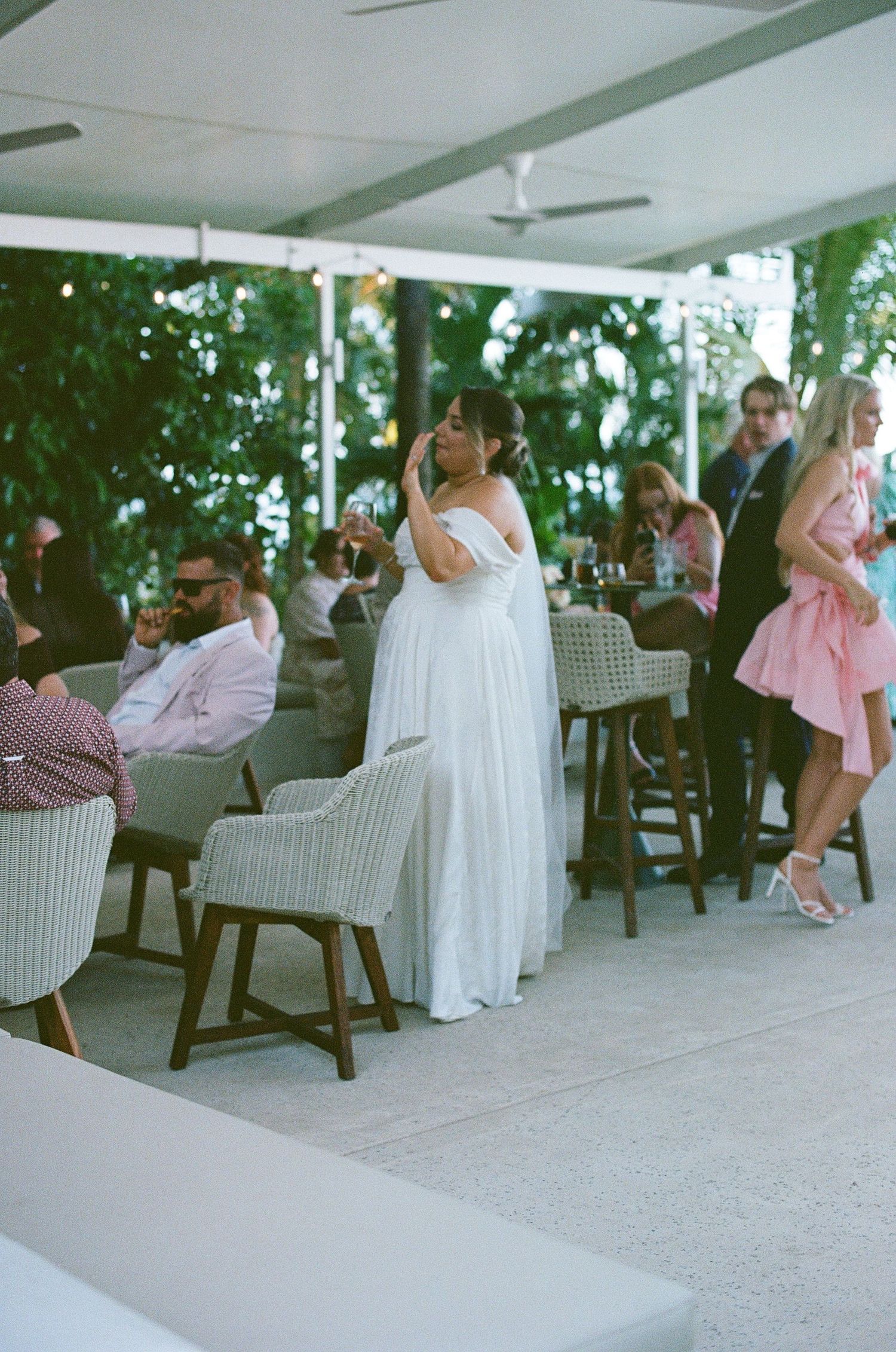 Bride in flowing white gown mingles with guests at an elegant open-air tropical wedding reception venue.