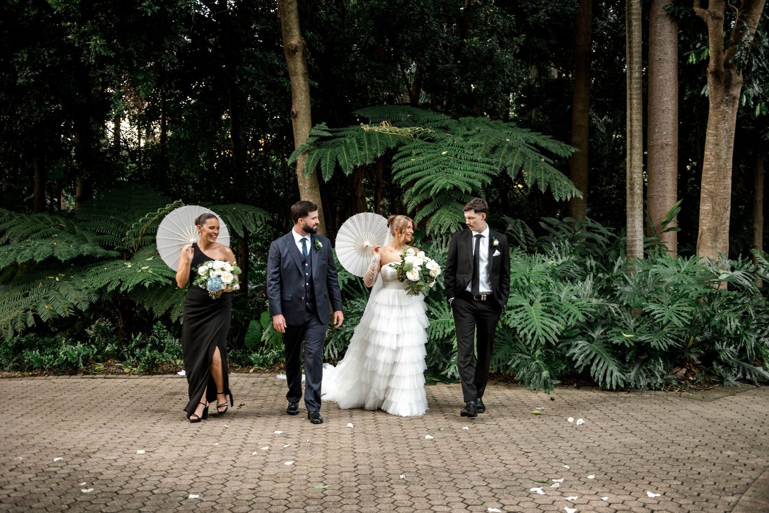 Wedding party walks through lush garden path, bridesmaids in black holding white parasols with bouquets.