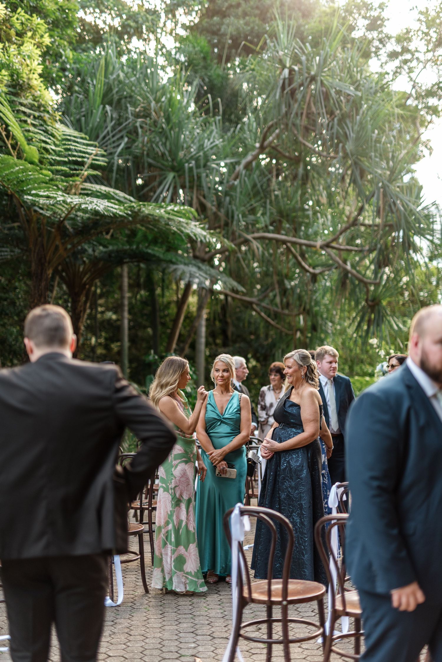 Wedding guests in formal attire mingling along a garden aisle lined with wooden chairs after the ceremony.