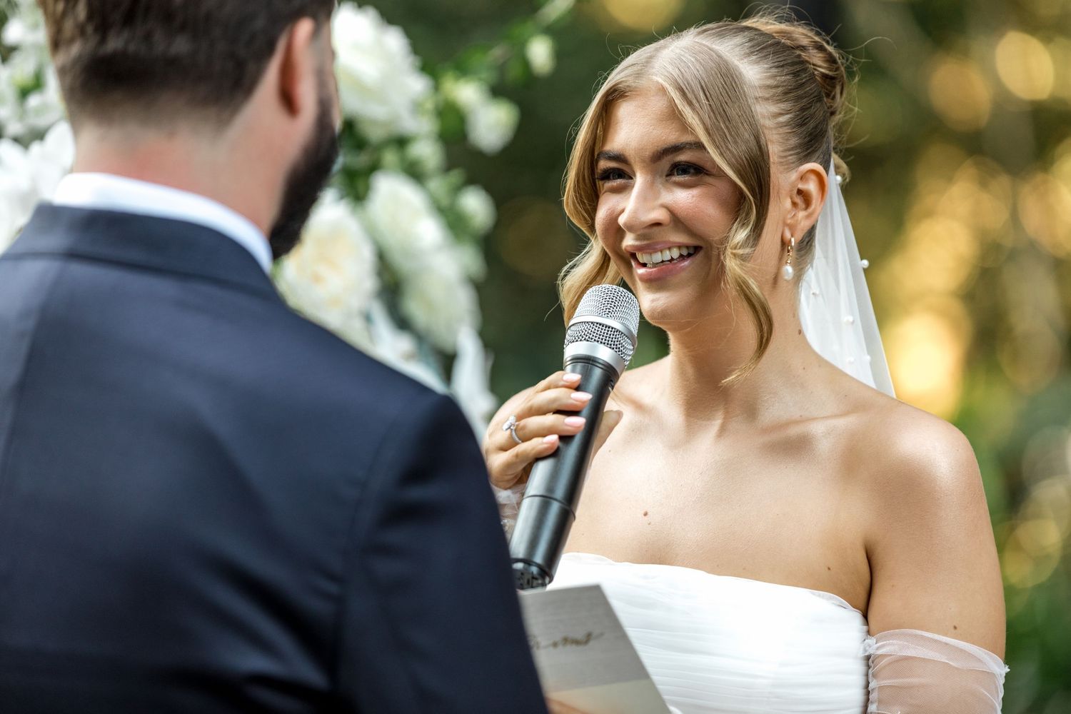 Radiant bride smiling while speaking vows into microphone during outdoor wedding ceremony.