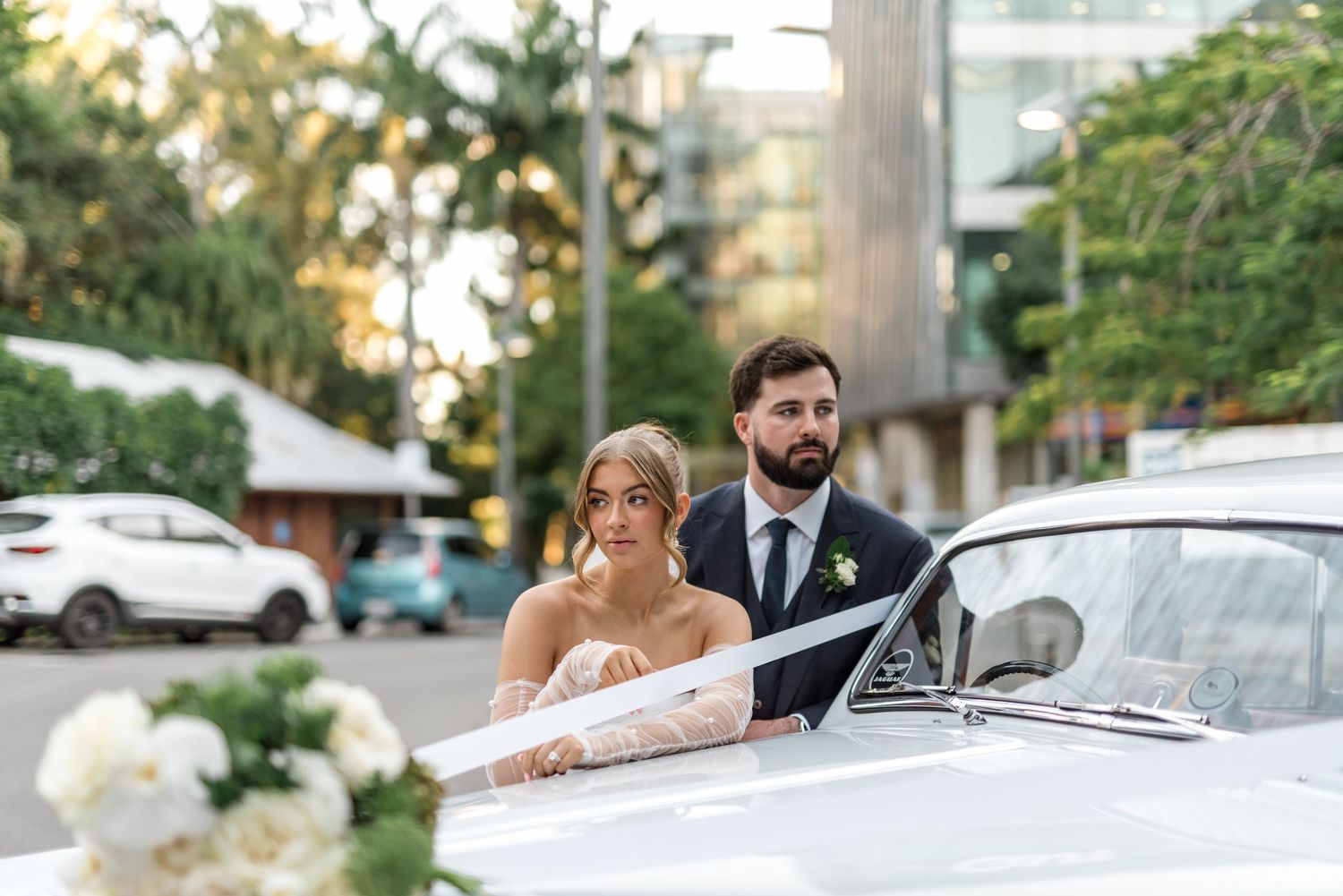 Bride and groom pose beside vintage white classic car decorated with white flowers at sunset.
