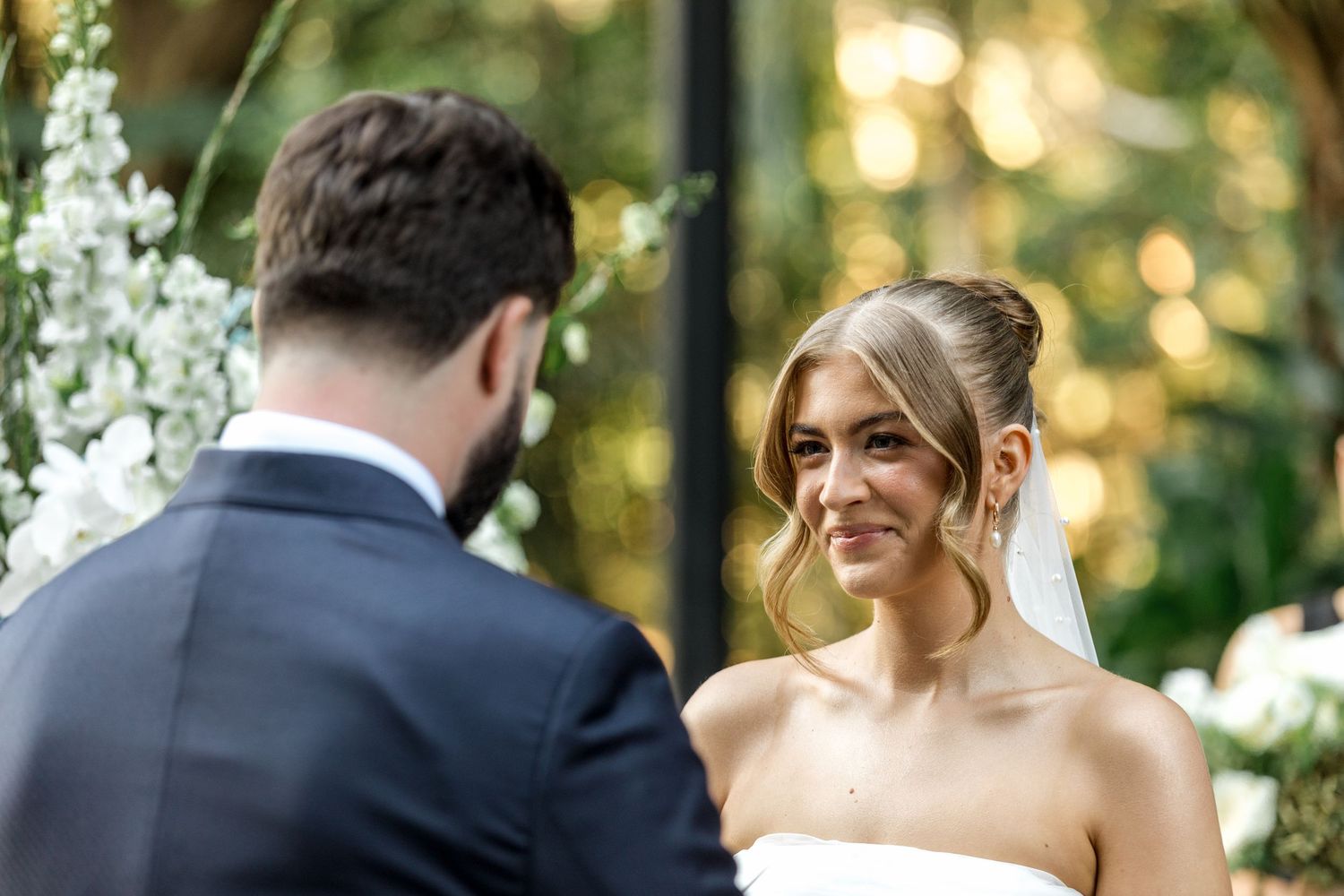 Bride gazes lovingly at groom during outdoor wedding ceremony with white floral arch backdrop.