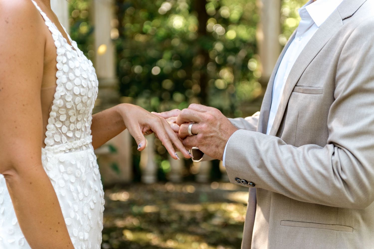 Groom slides wedding band onto brides finger during outdoor ceremony, both wearing elegant wedding attire.