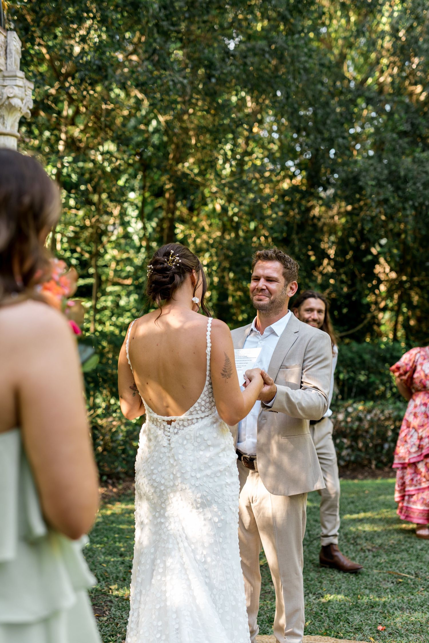 Groom smiling joyfully at bride in backless lace gown during outdoor garden wedding ceremony.
