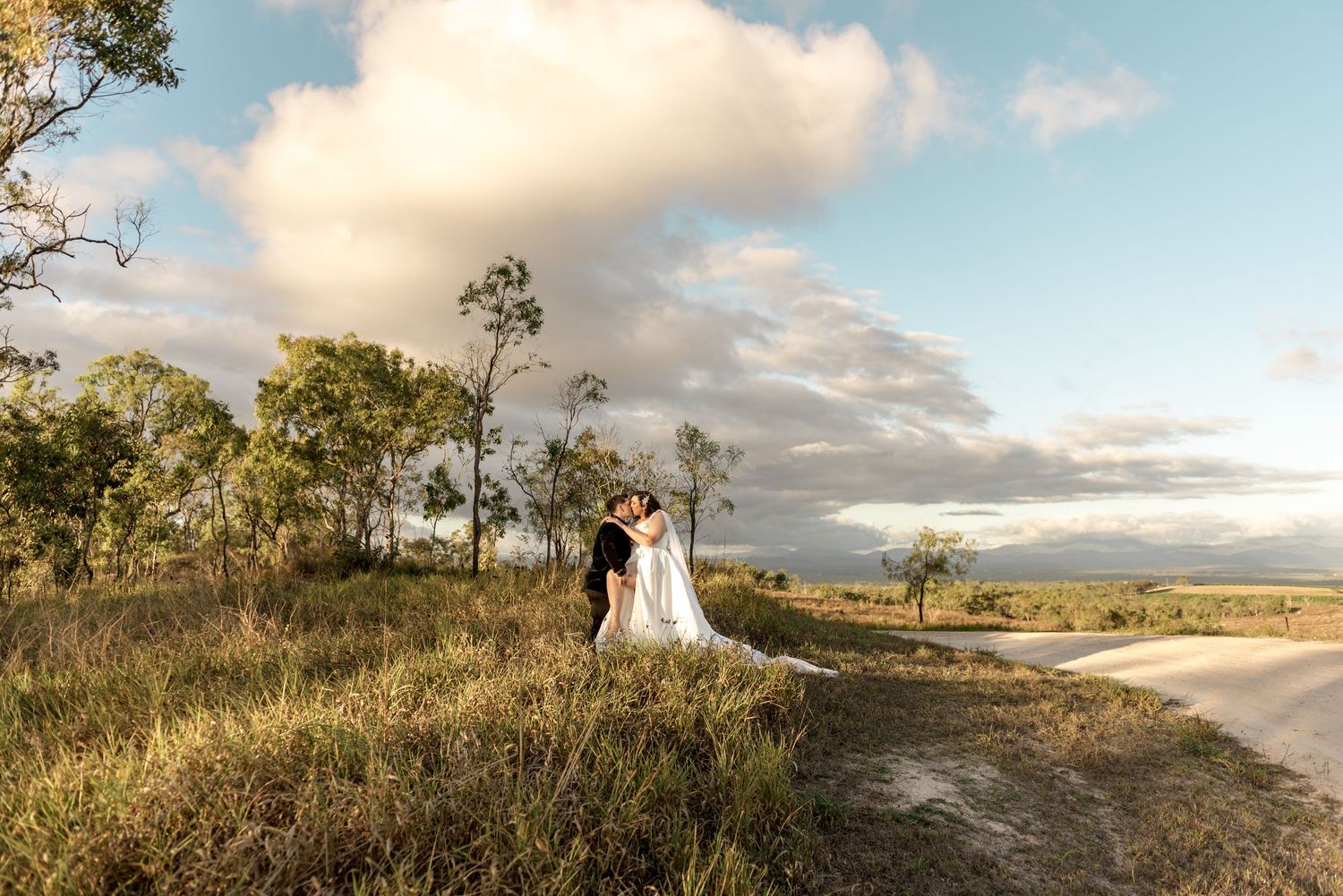 Bride and groom kissing on a scenic rural road surrounded by golden landscape.