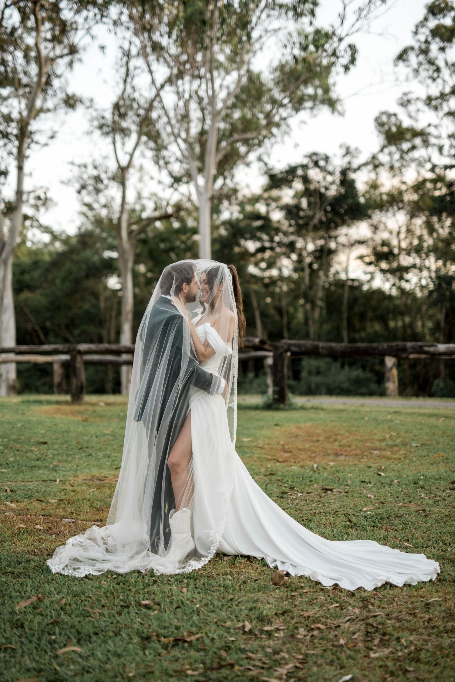 Bride and groom sharing romantic kiss under flowing white bridal veil in lush green outdoor setting.