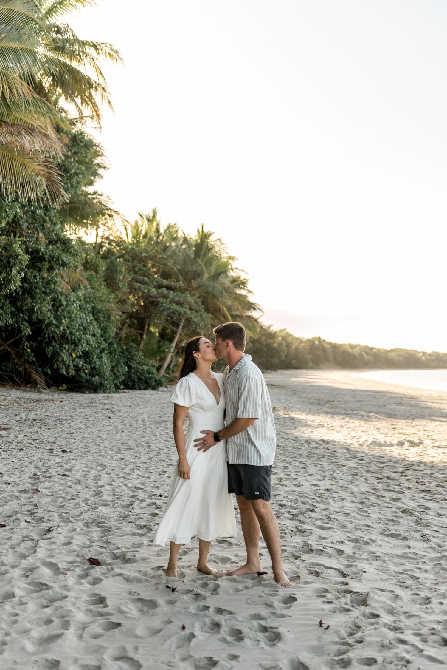 Couple sharing a romantic moment on a sunny tropical beach surrounded by lush green palm trees.