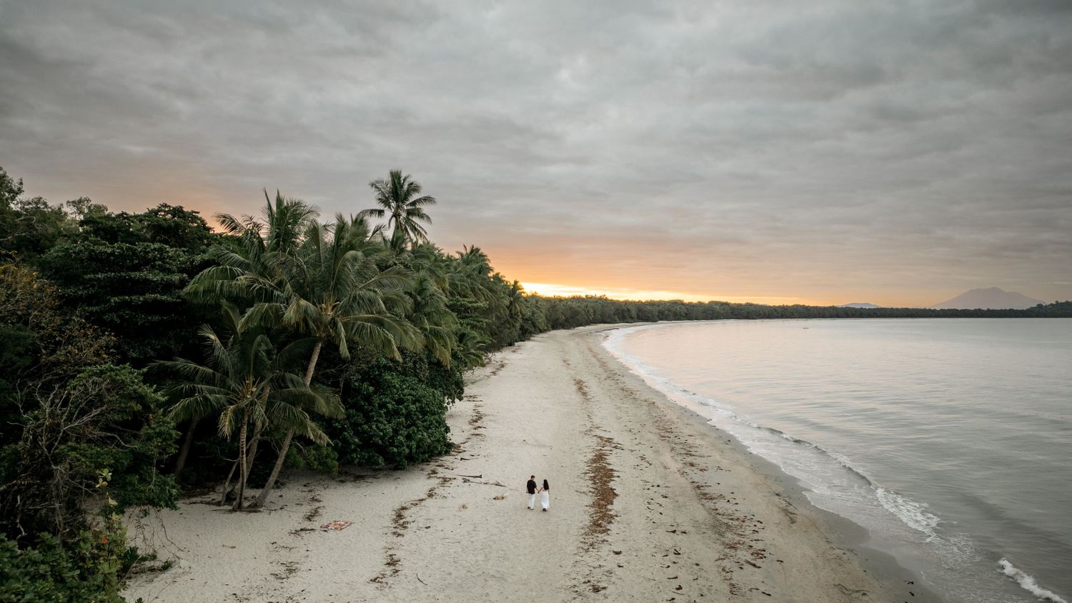 Aerial view of a tropical beach at sunset with palm trees and two tiny figures on the sand.