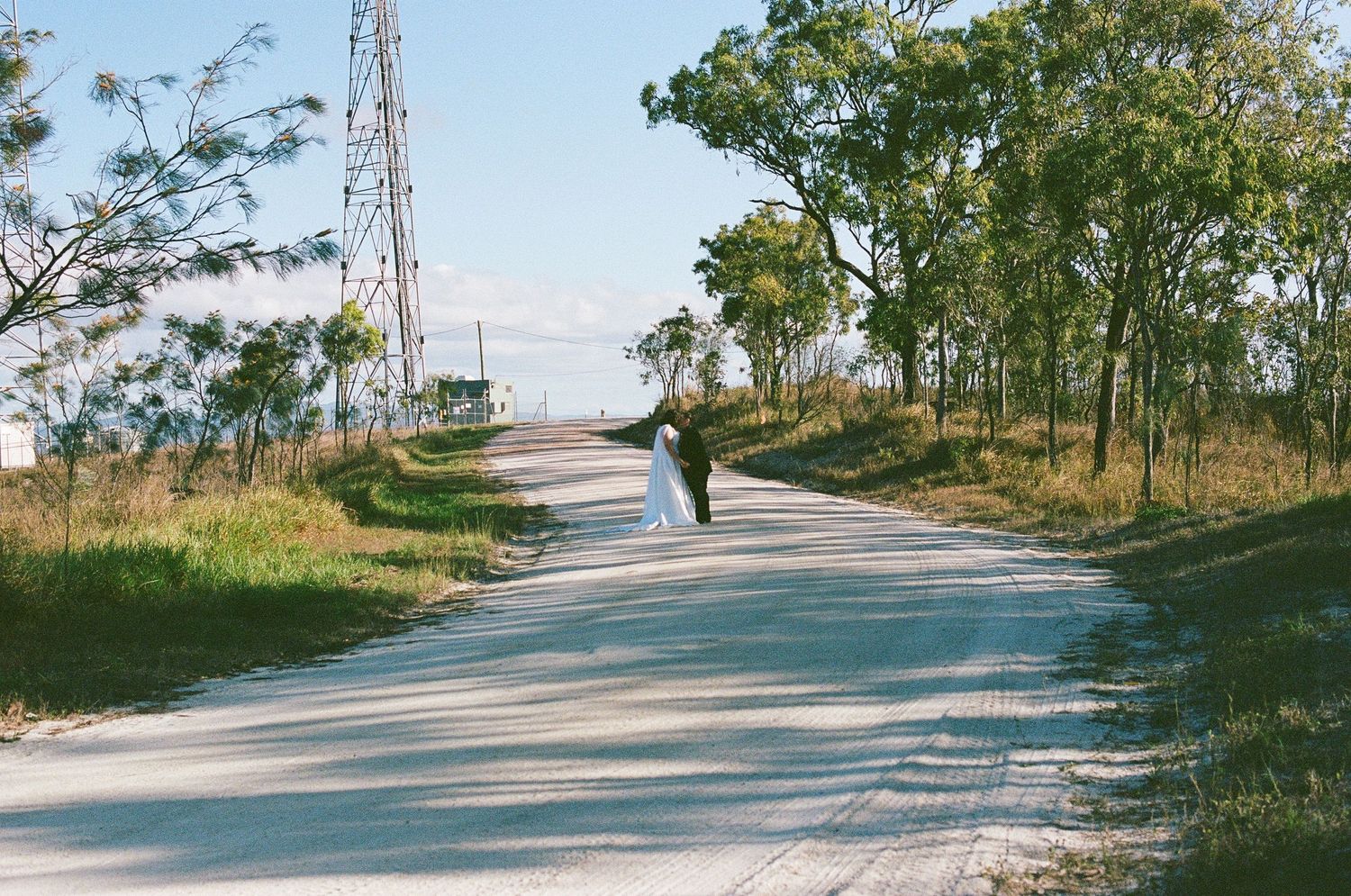 Bride and groom walk hand in hand down a tree-lined rural road on their wedding day.