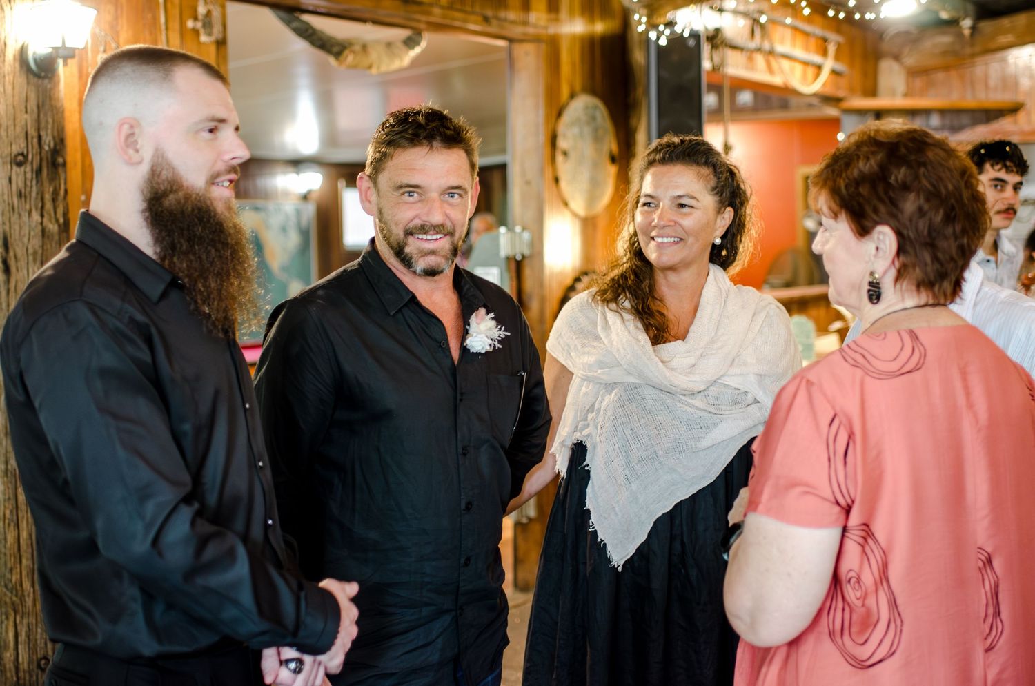 Two men in black suits smile and chat with bride and guests at a rustic wedding reception.