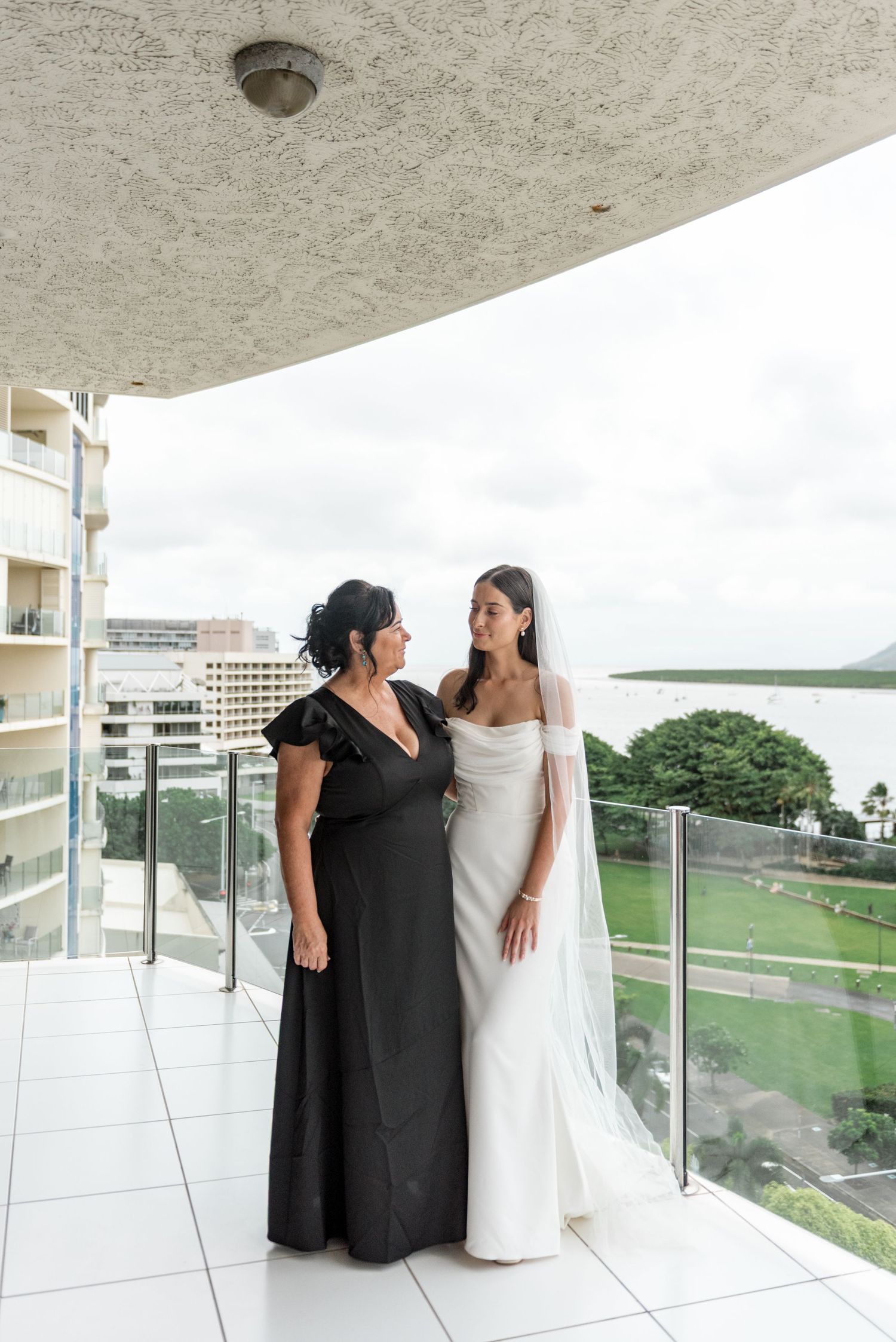 Bride in white off-shoulder gown and veil stands with mother in black dress on balcony with ocean view.