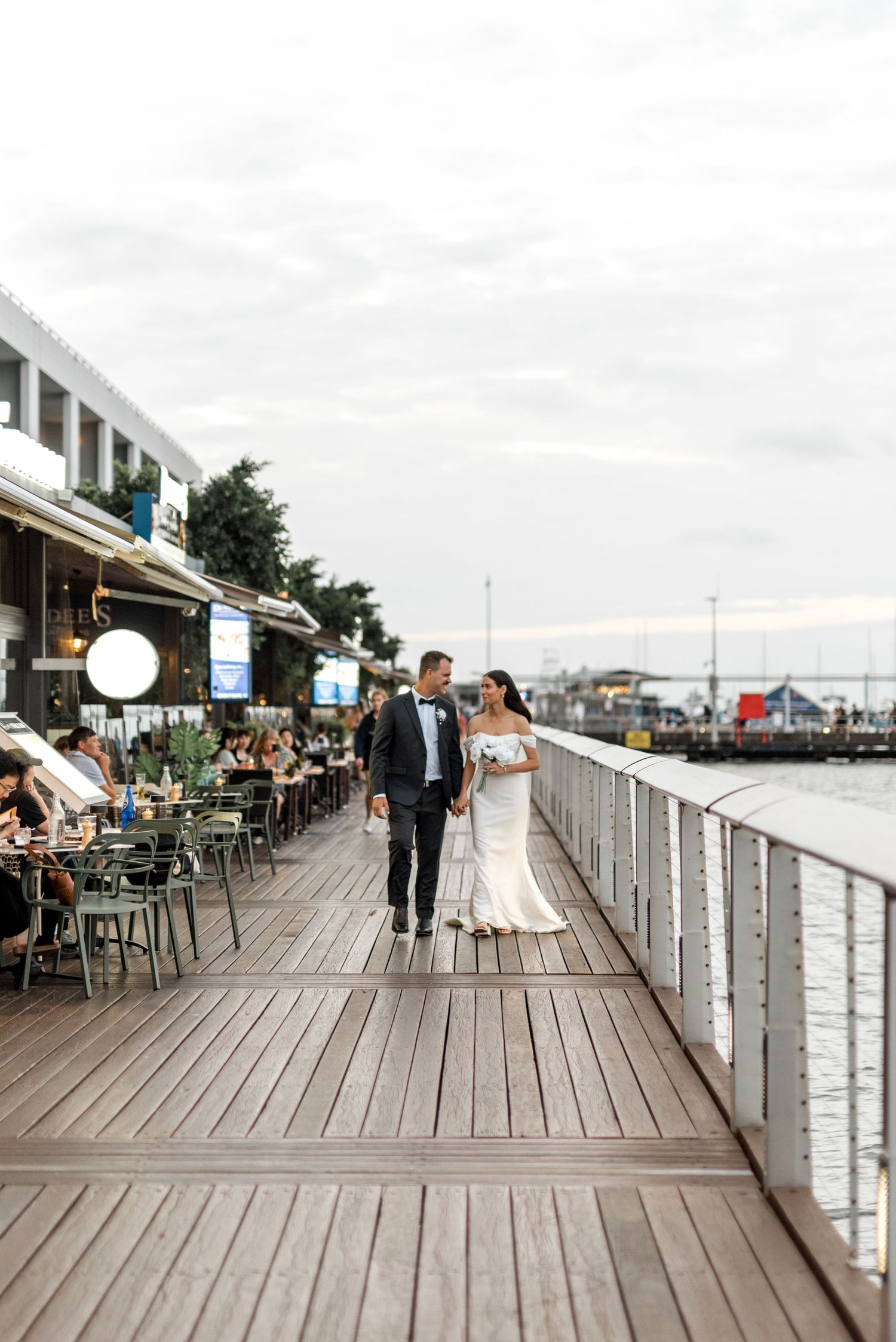 Bride and groom stroll along a waterfront boardwalk with harbor views and dining guests nearby.