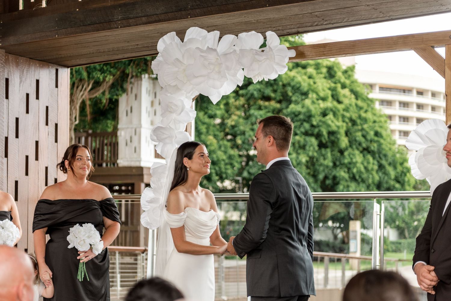 Bride and groom exchange vows under white floral arch at outdoor wedding ceremony.