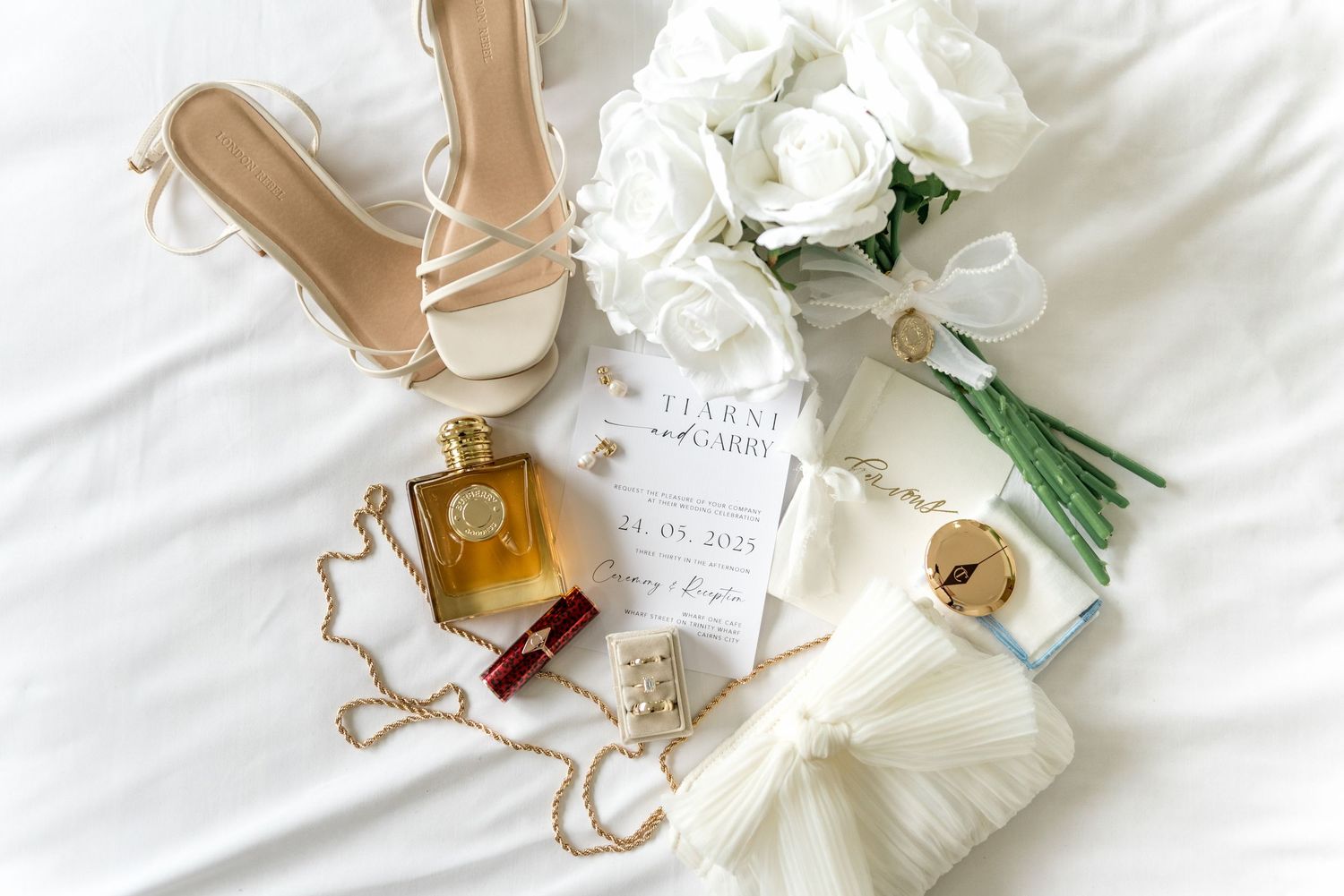 Elegant bridal flat lay with white roses, nude heels, gold perfume, red lipstick, pearl necklace, and wedding invitation.