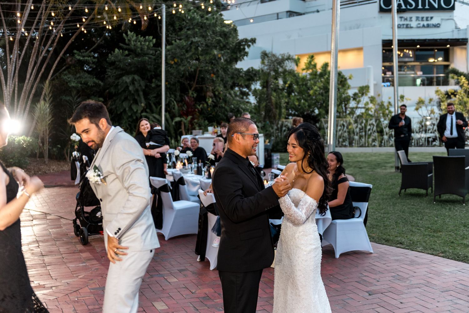 Wedding guests join the bride and groom on the dance floor during a lively outdoor evening reception celebration.