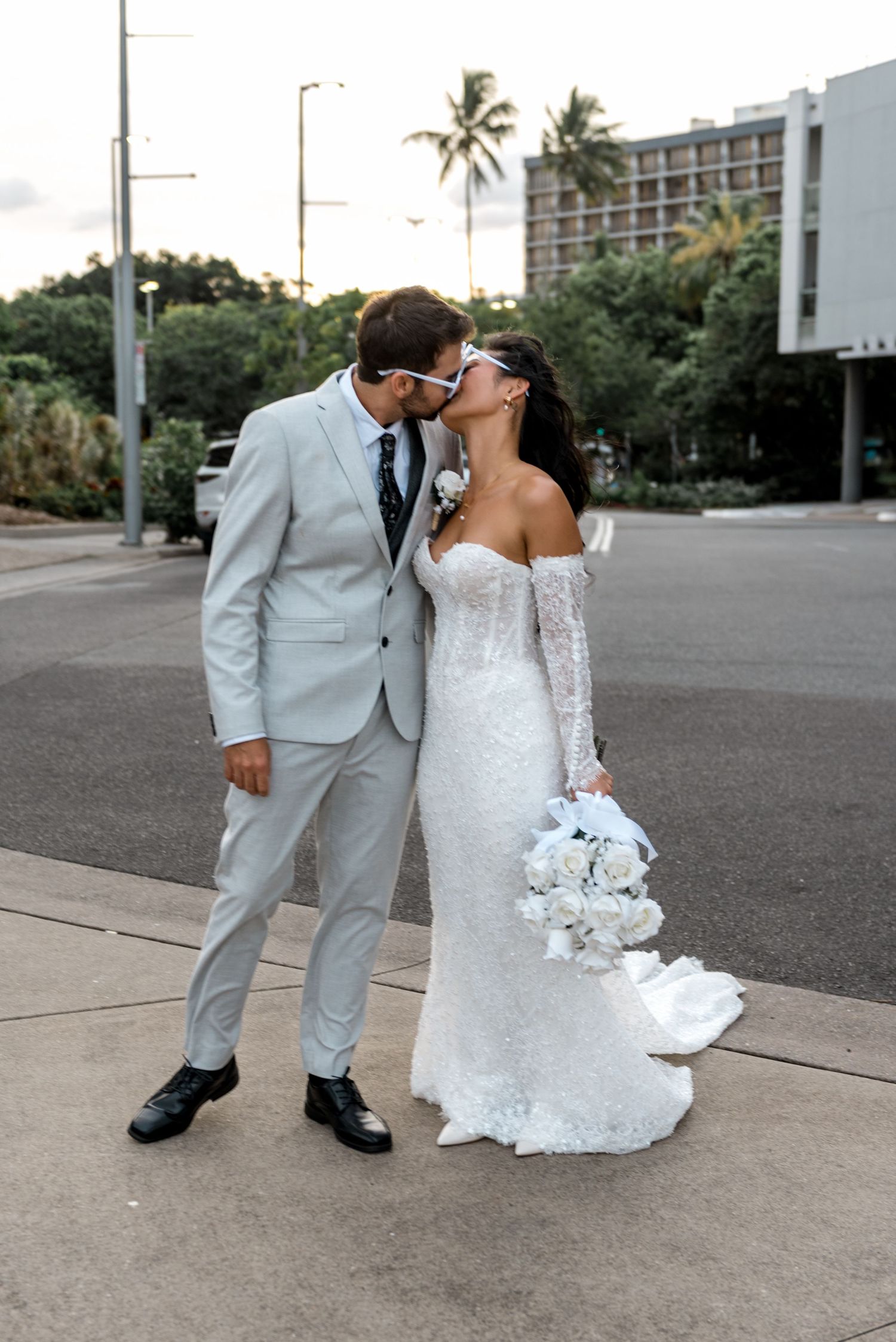 Bride and groom sharing a kiss on sidewalk wearing novelty sunglasses with palm trees and hotel in background.