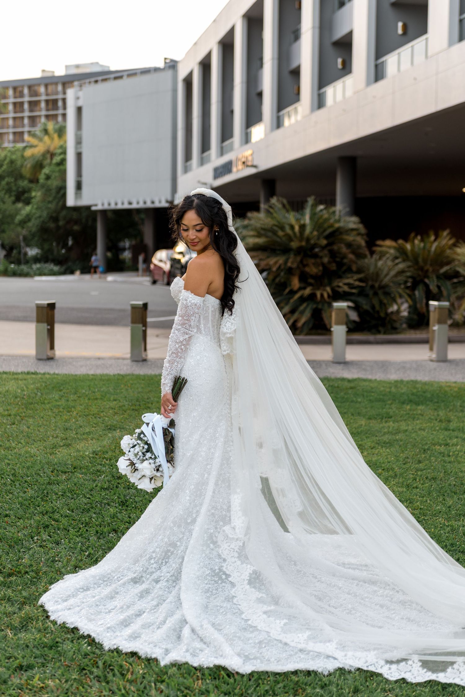 Bride in long-sleeve lace mermaid gown with cathedral veil posing on lawn in front of modern building.