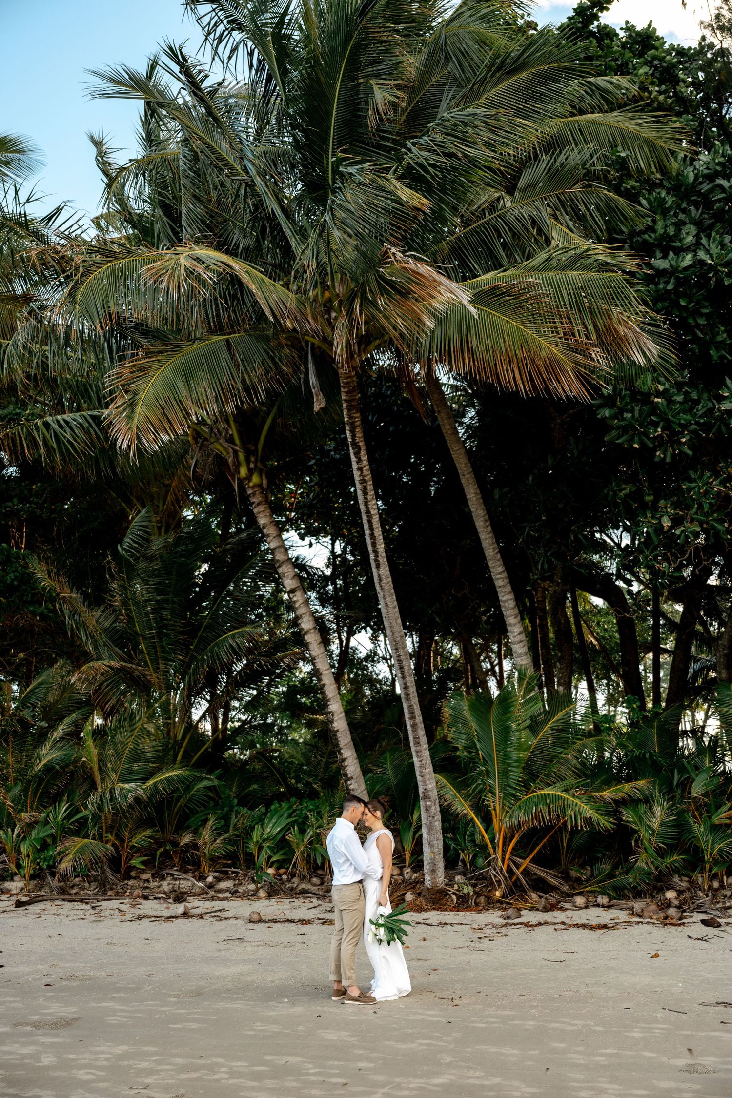 Newlyweds share a kiss beneath tall swaying palm trees on a tropical sandy beach at dusk at Four Mile Beach, Port Douglas.