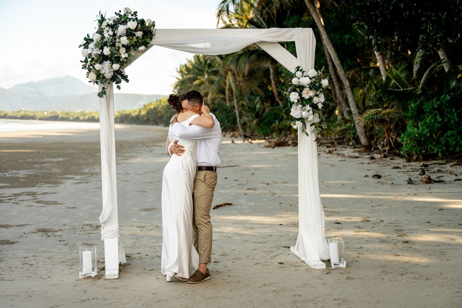 Newlyweds embracing in a romantic hug under a white floral arch on a tropical sandy beach after their ceremony at Four Mile Beach, Port Dou.