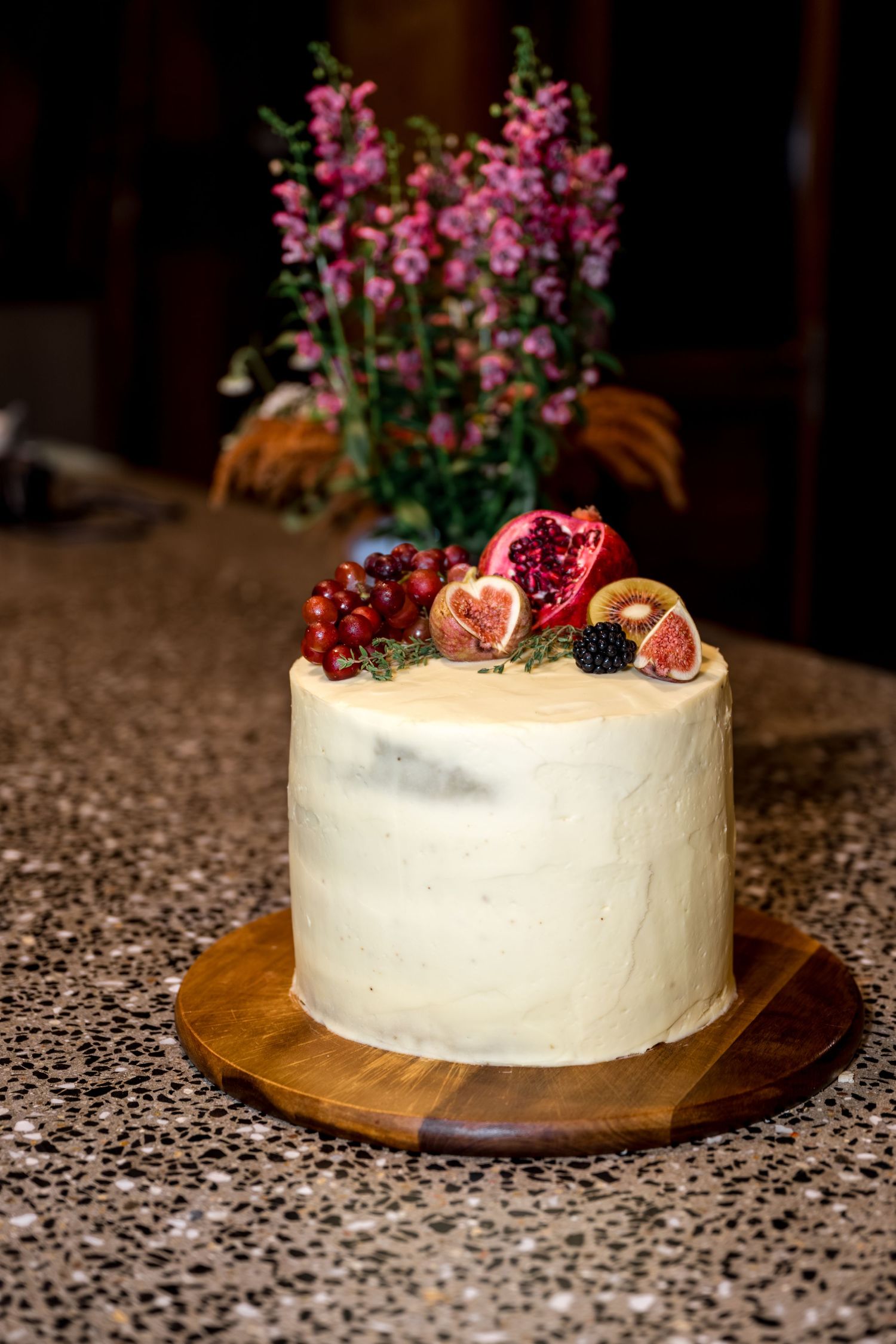 Elegant white single-tier cake topped with fresh figs, pomegranate, and berries on a wooden board.