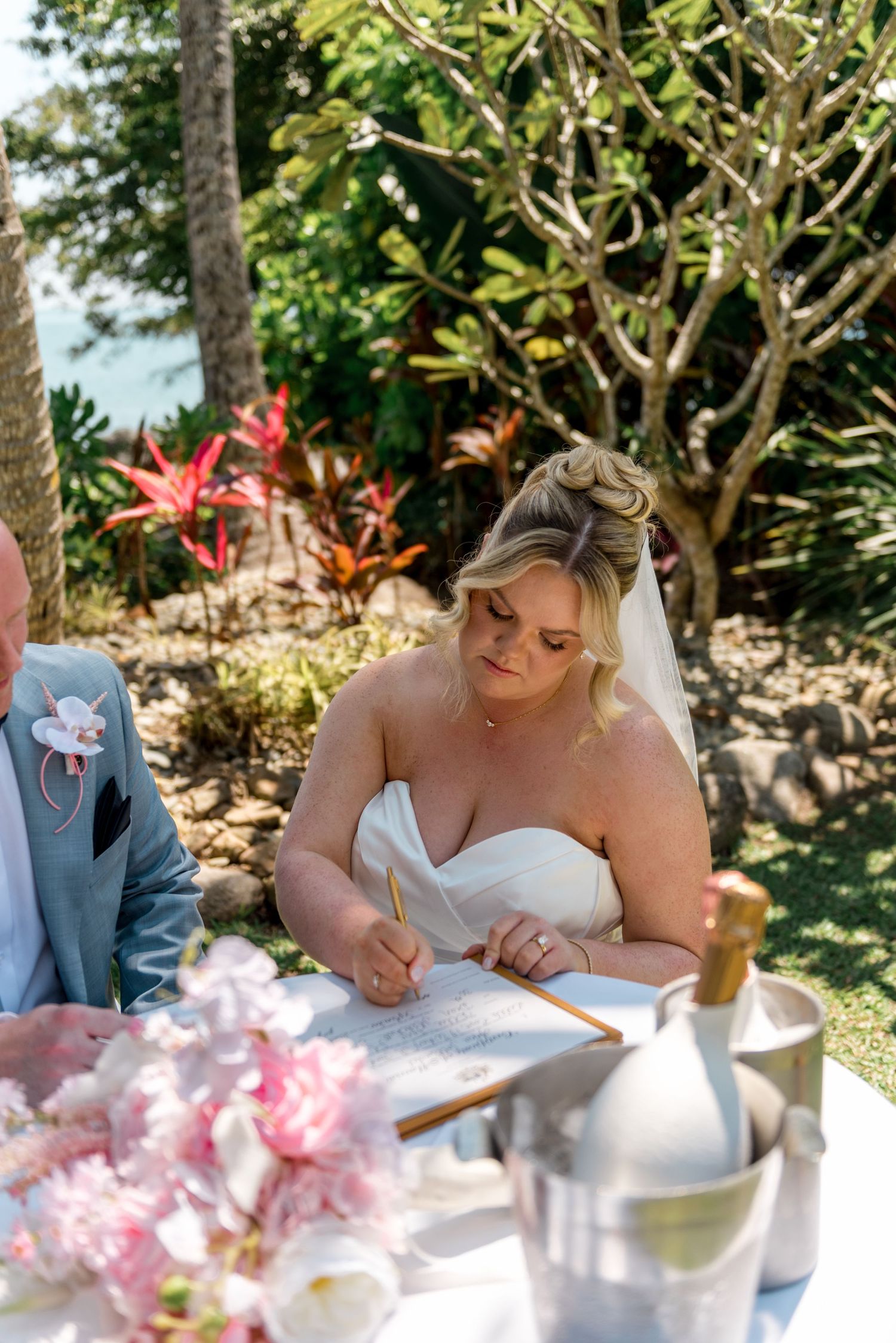 Blonde bride in strapless white gown signing wedding certificate with golden pen at outdoor ceremony table.