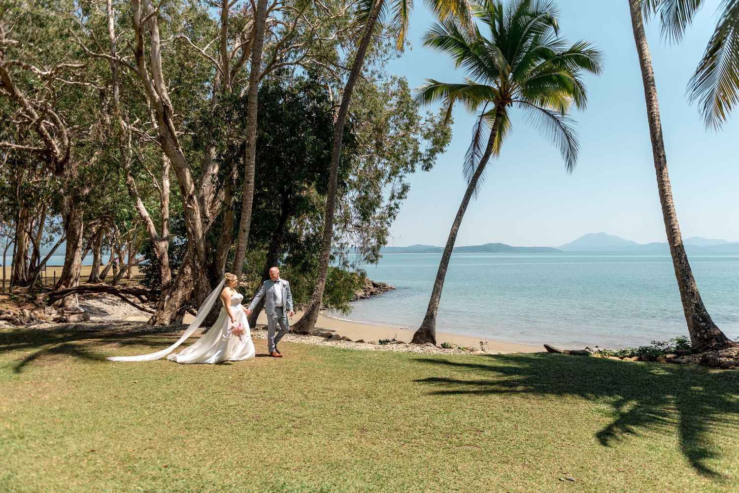 Newlyweds walk hand in hand along a lush tropical beachfront lawn lined with palm trees and ocean views.