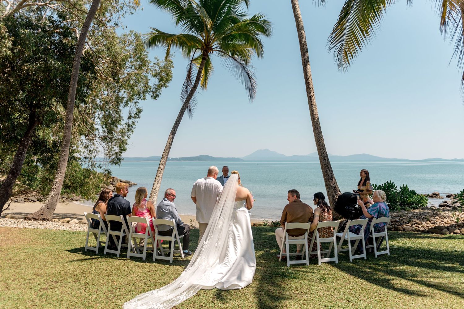 Bride with long veil walks down aisle at beachside wedding ceremony under palm trees.