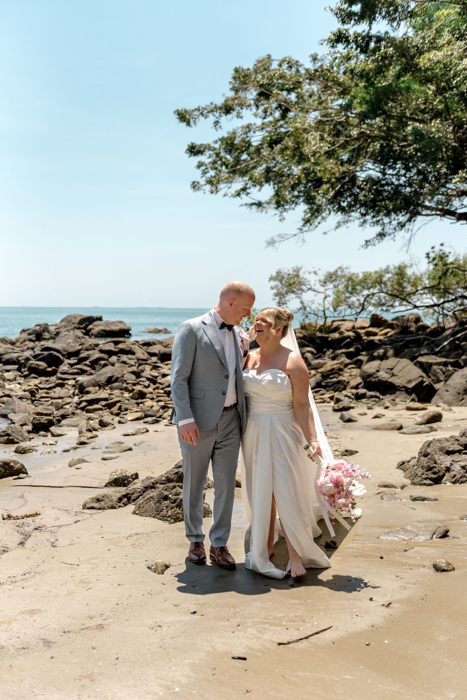Bride holding pink bouquet and groom in grey suit sharing a tender moment on sandy beach.
