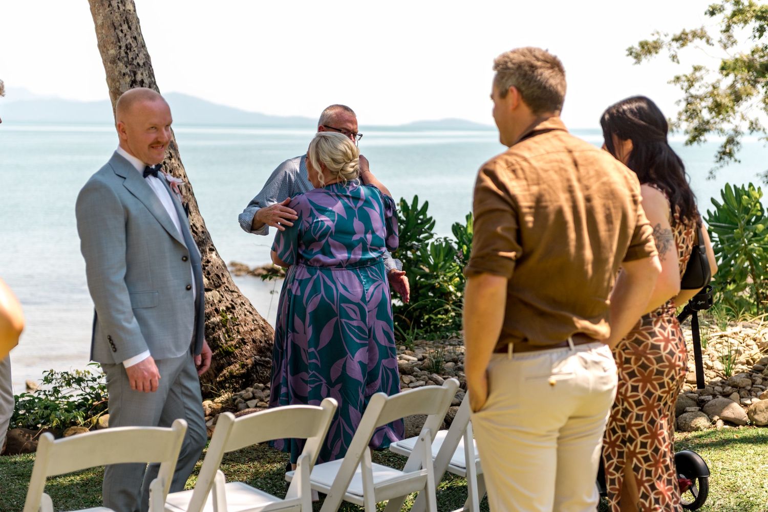 Wedding guests in colorful attire chat near white chairs at a tropical beachside ceremony.