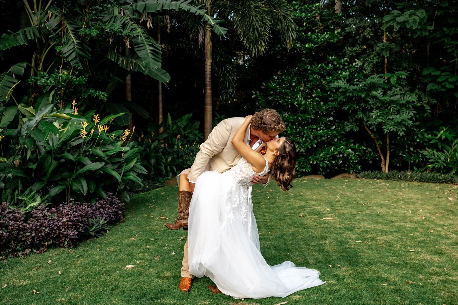 Groom dipping bride in a romantic kiss amid lush tropical garden greenery.