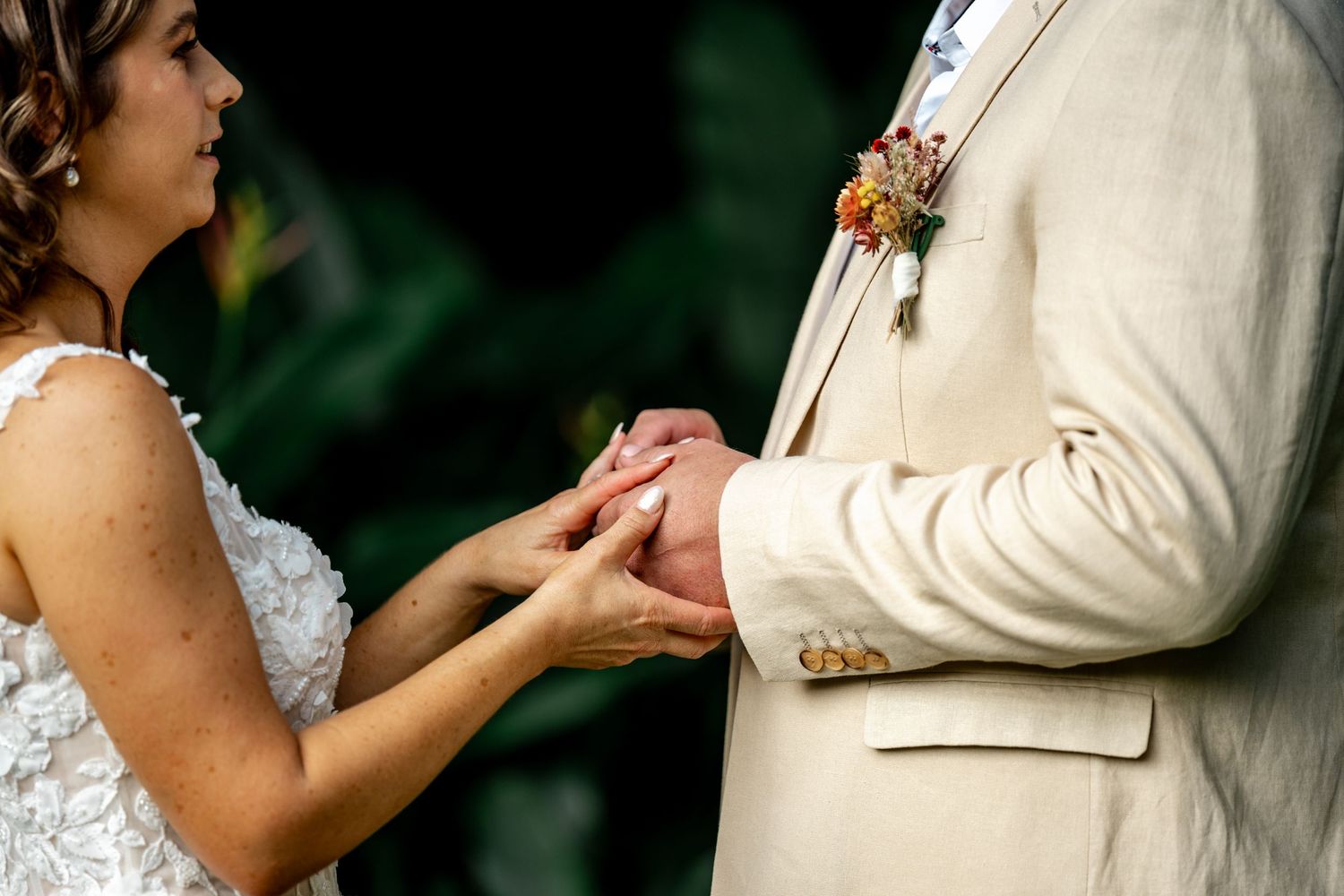 Bride placing wedding ring on groom's finger during outdoor ceremony, groom wearing beige suit with floral boutonniere.