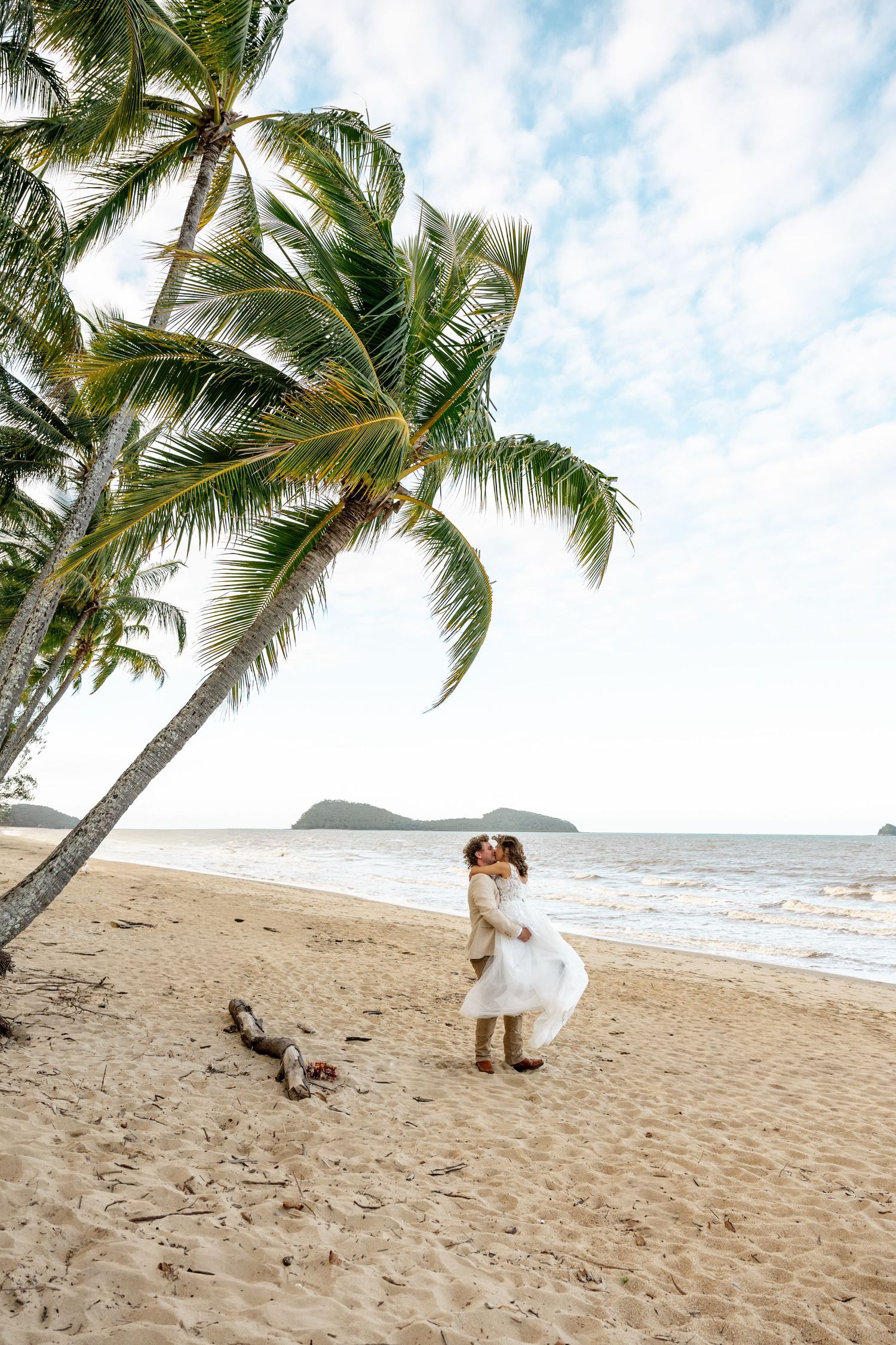 Groom lifts bride in a joyful embrace under a palm tree on a golden tropical sandy beach.