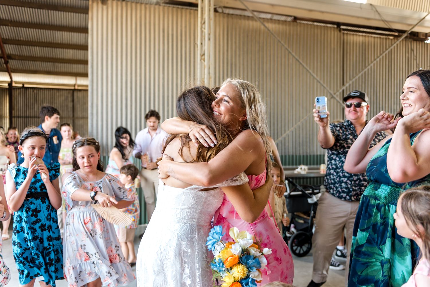 Bride hugs smiling guest holding colorful bouquet at lively outdoor barn wedding.