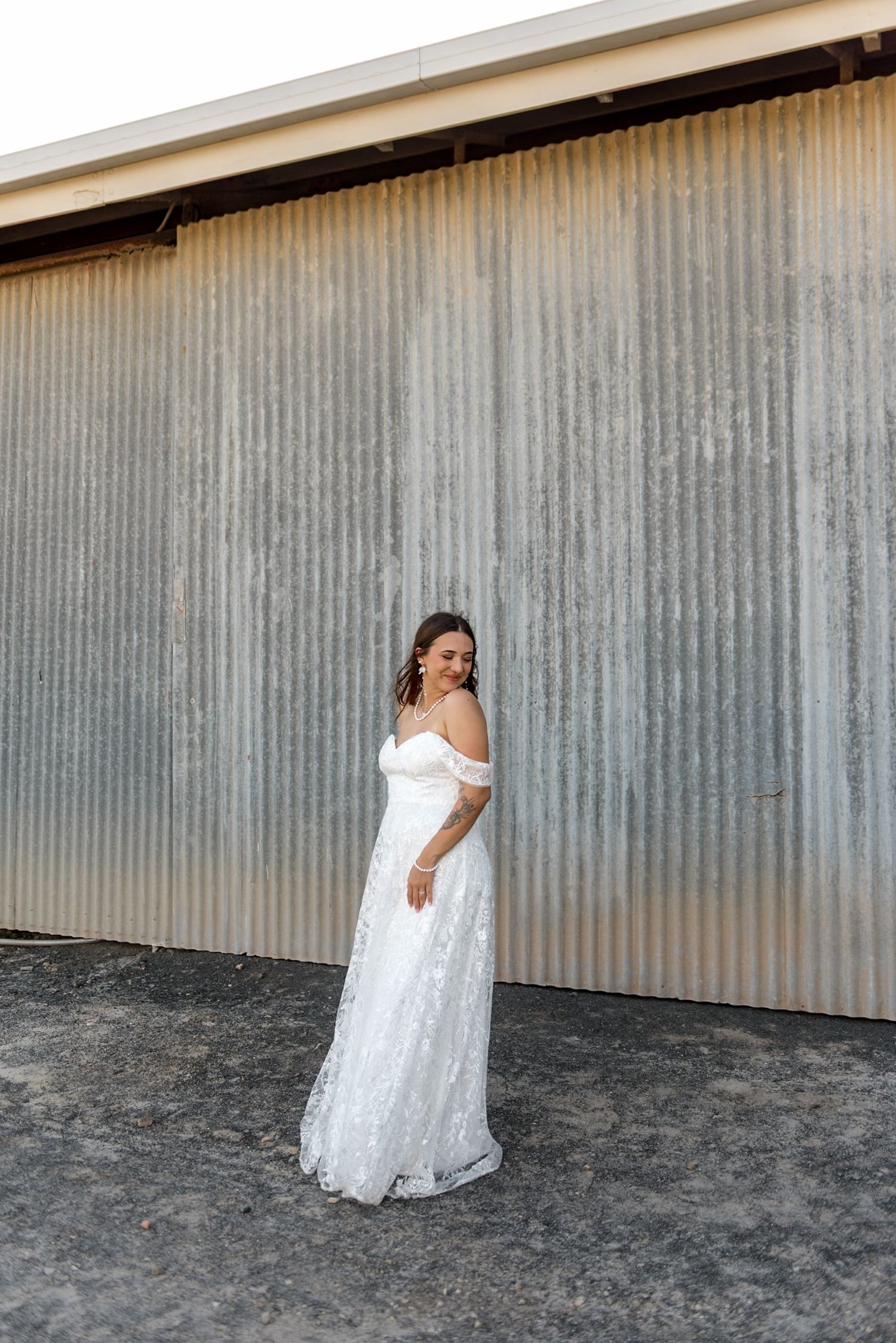 Bride in off-shoulder lace wedding gown poses elegantly in front of rustic corrugated metal barn wall.