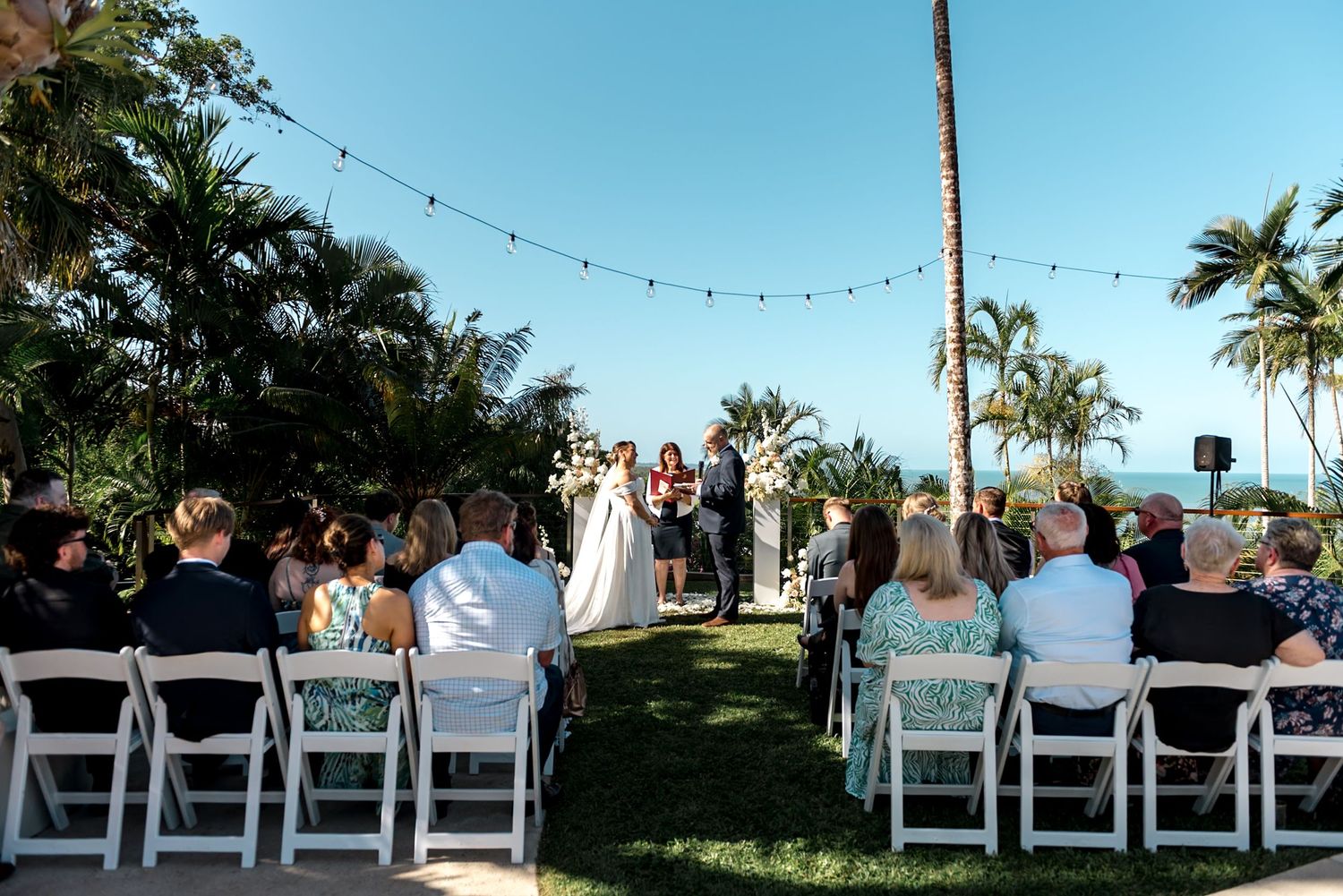 Outdoor tropical wedding ceremony with guests seated in white chairs facing altar decorated with white florals.