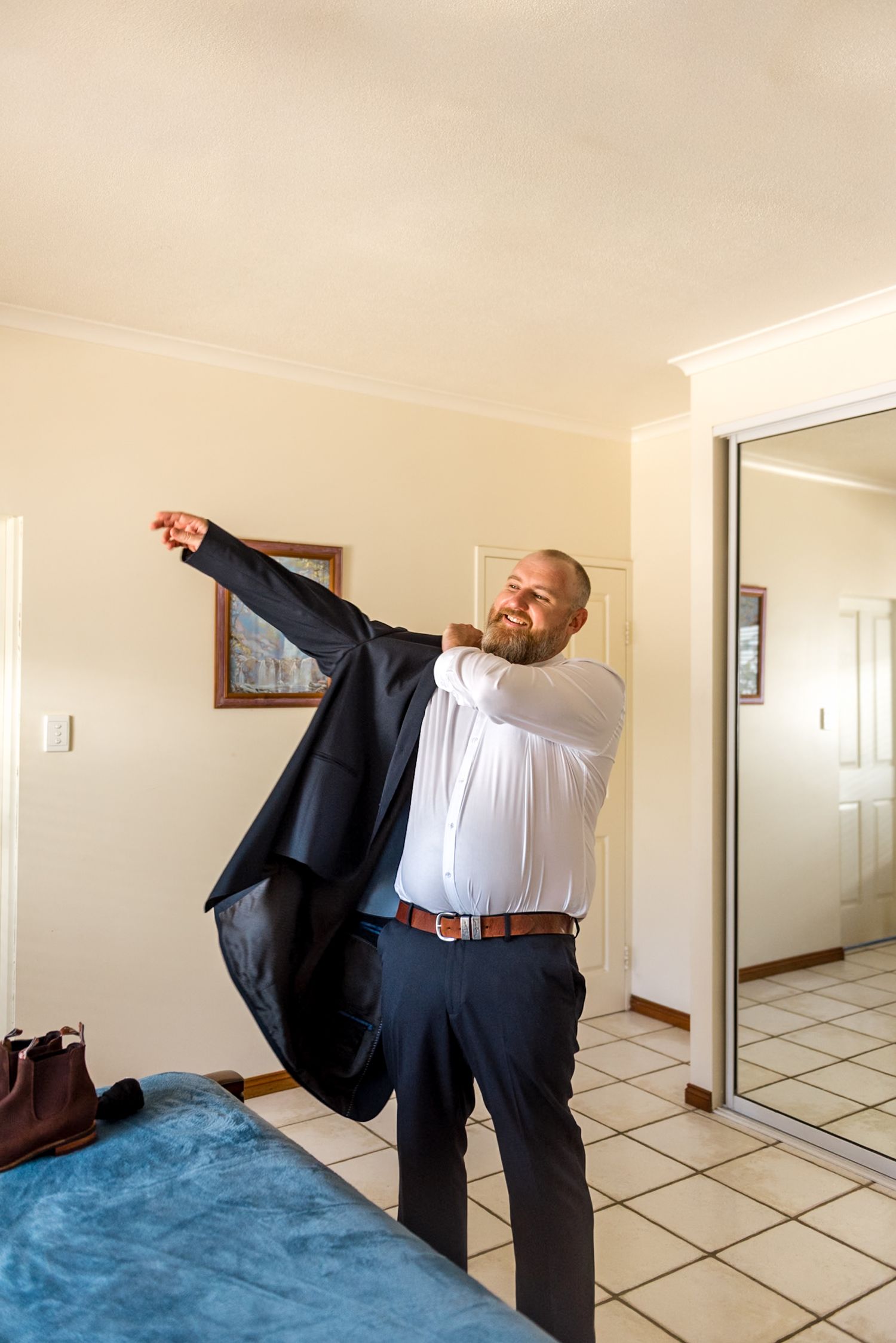 Bearded groom putting on dark navy suit jacket while getting ready for wedding in bedroom.