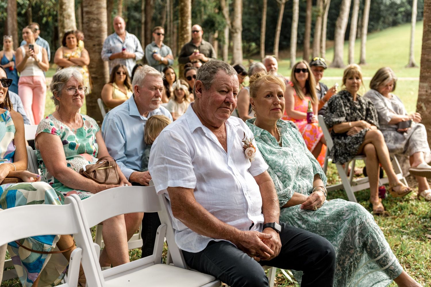 Wedding guests seated on white chairs outdoors watching ceremony in tropical garden setting.
