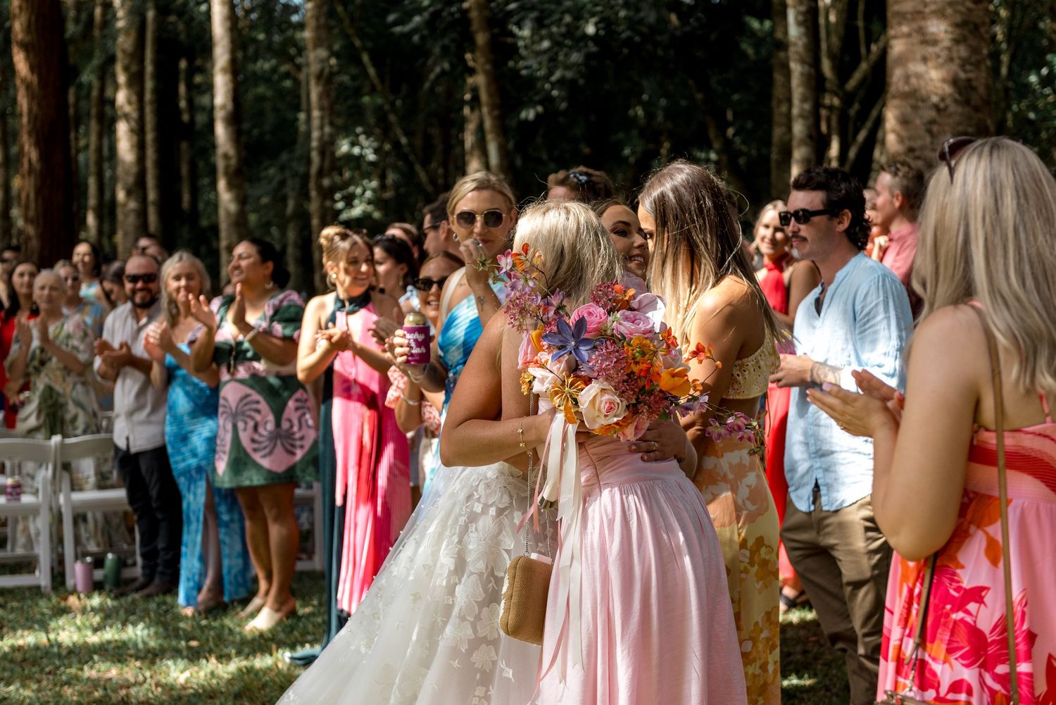Wedding guests in colorful attire applaud as bride holds vibrant bouquet during outdoor forest wedding celebration.