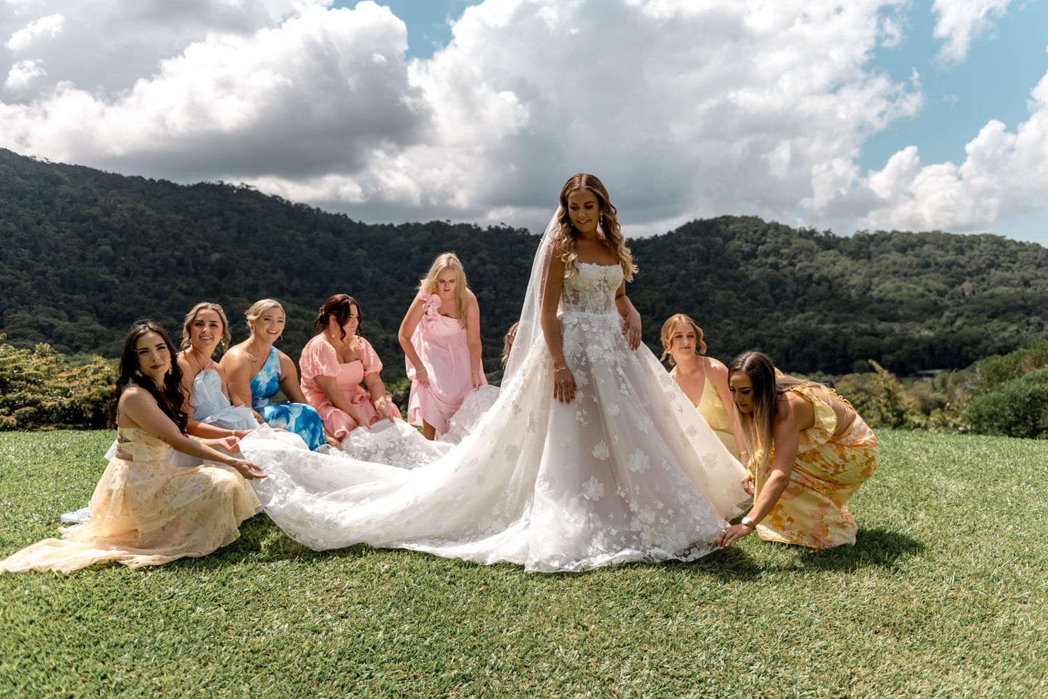 Bride in floral lace gown poses with colorful bridesmaids on green lawn with mountain backdrop.
